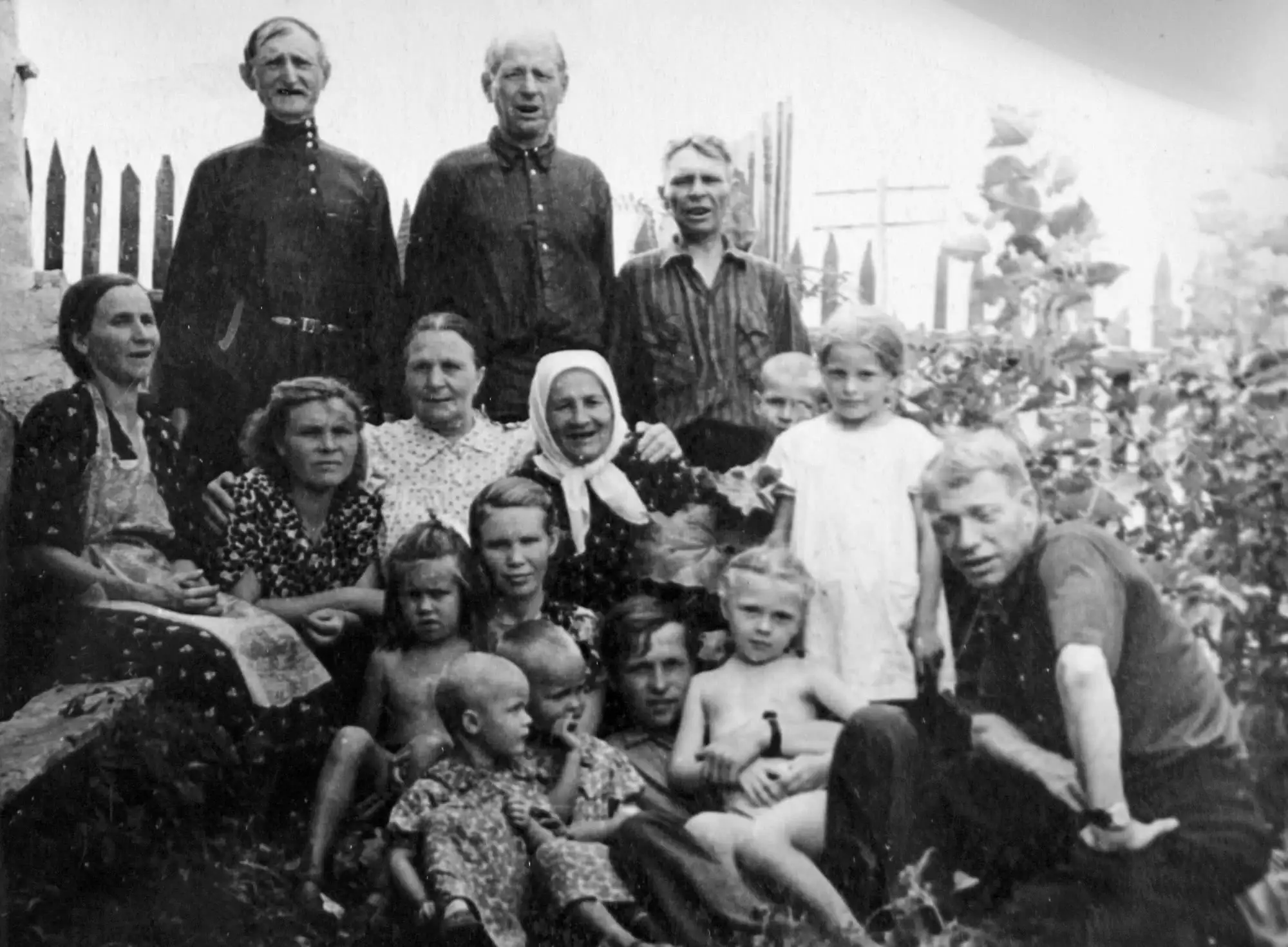 Чёрно-белая фотография большой семьи: мужчины, женщины и дети разных возрастов позируют на улице у забора и кустов, некоторые сидят, другие стоят.