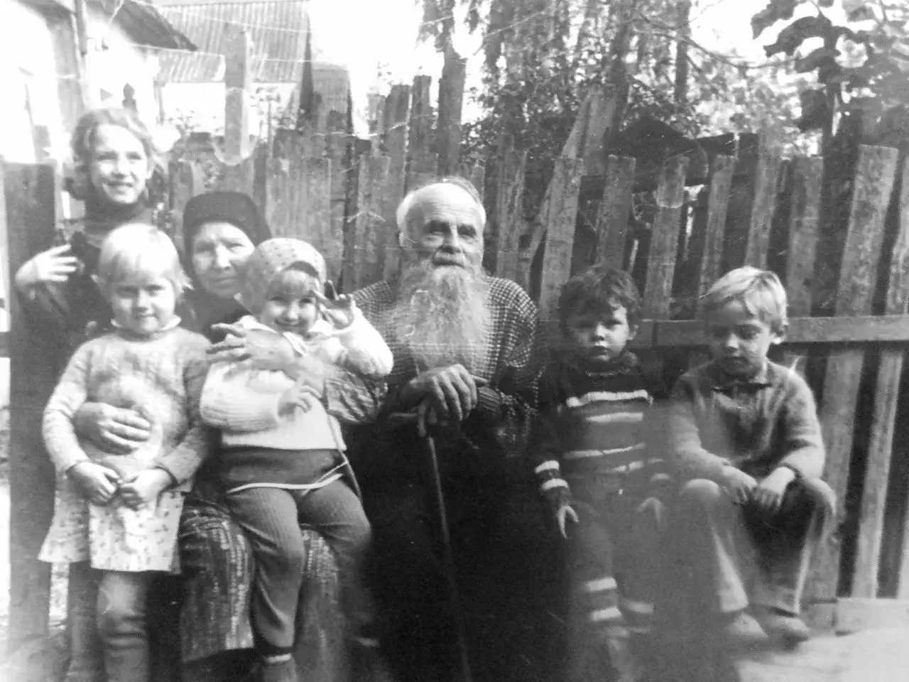 Чёрно-белое фото: пожилой мужчина с длинной бородой сидит в центре, окружённый шестью детьми разного возраста на фоне деревянного забора и деревьев.