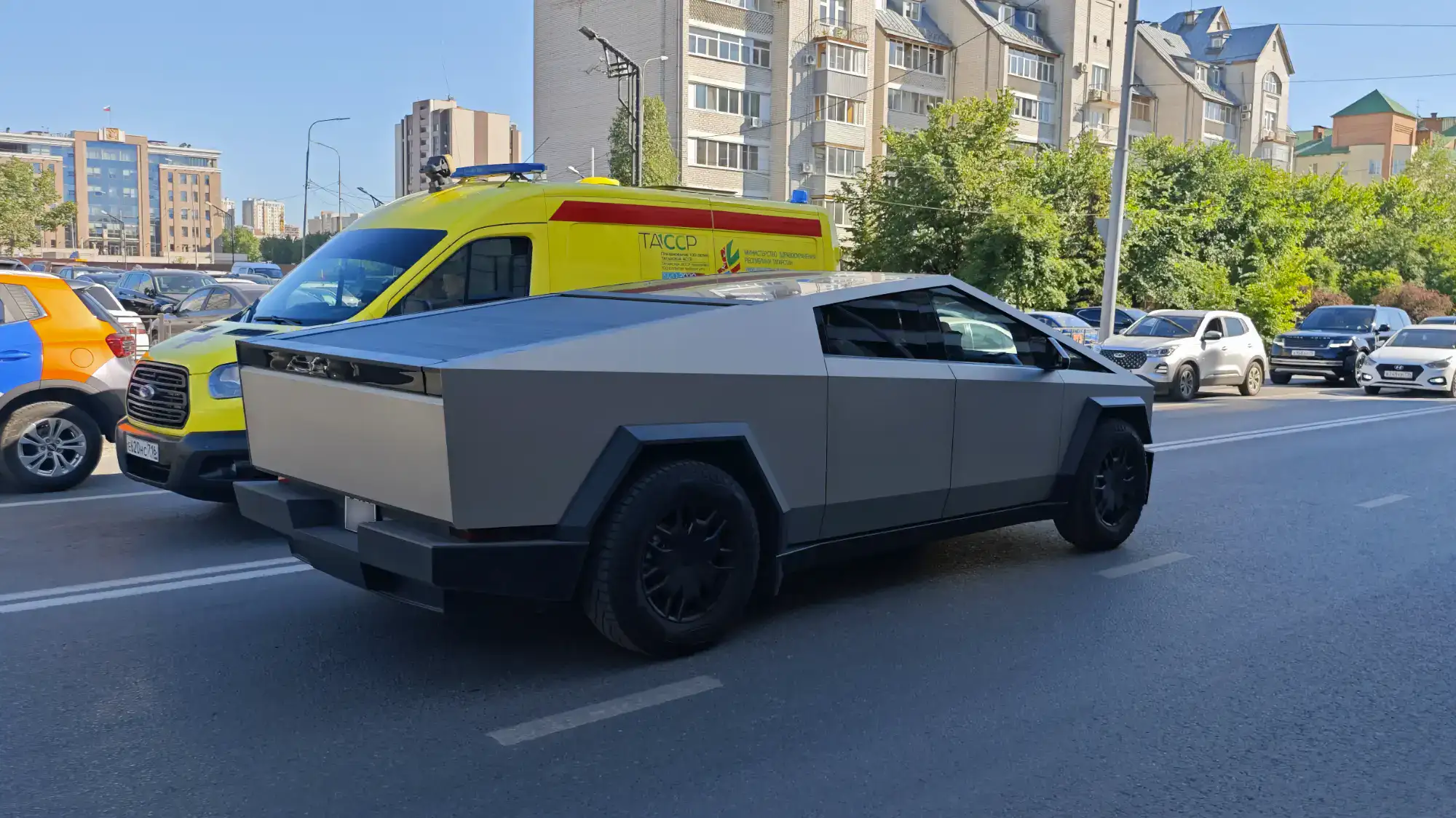 Серый пикап Tesla Cybertruck с угловатым дизайном едет по городской дороге, рядом с ним — желтая машина скорой помощи с надписью «ТАССР» и синими проблесковыми маячками.