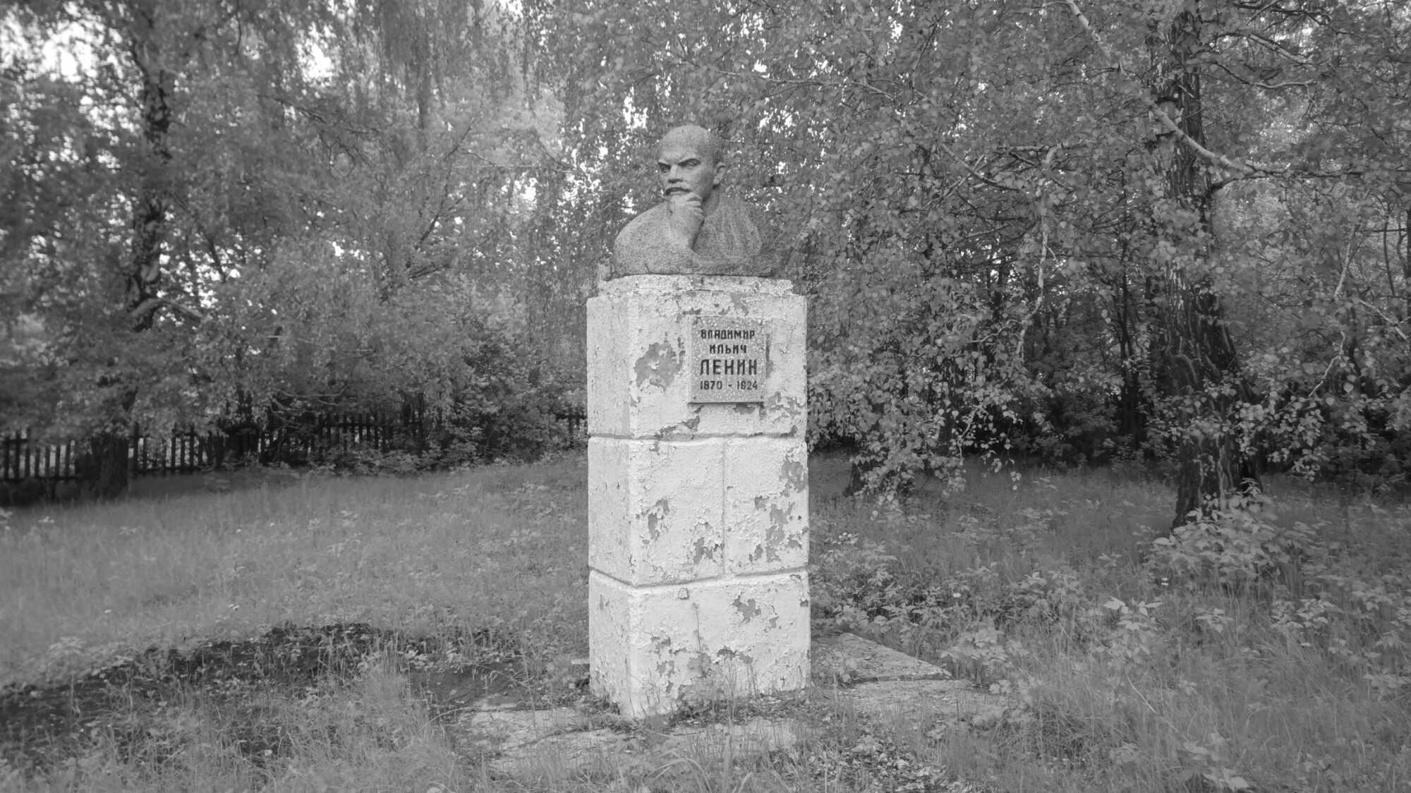 Бюст Ленина на постаменте с надписью «Владимир Ильич Ленин 1870–1924», окружённый деревьями и травой, чёрно-белое фото.