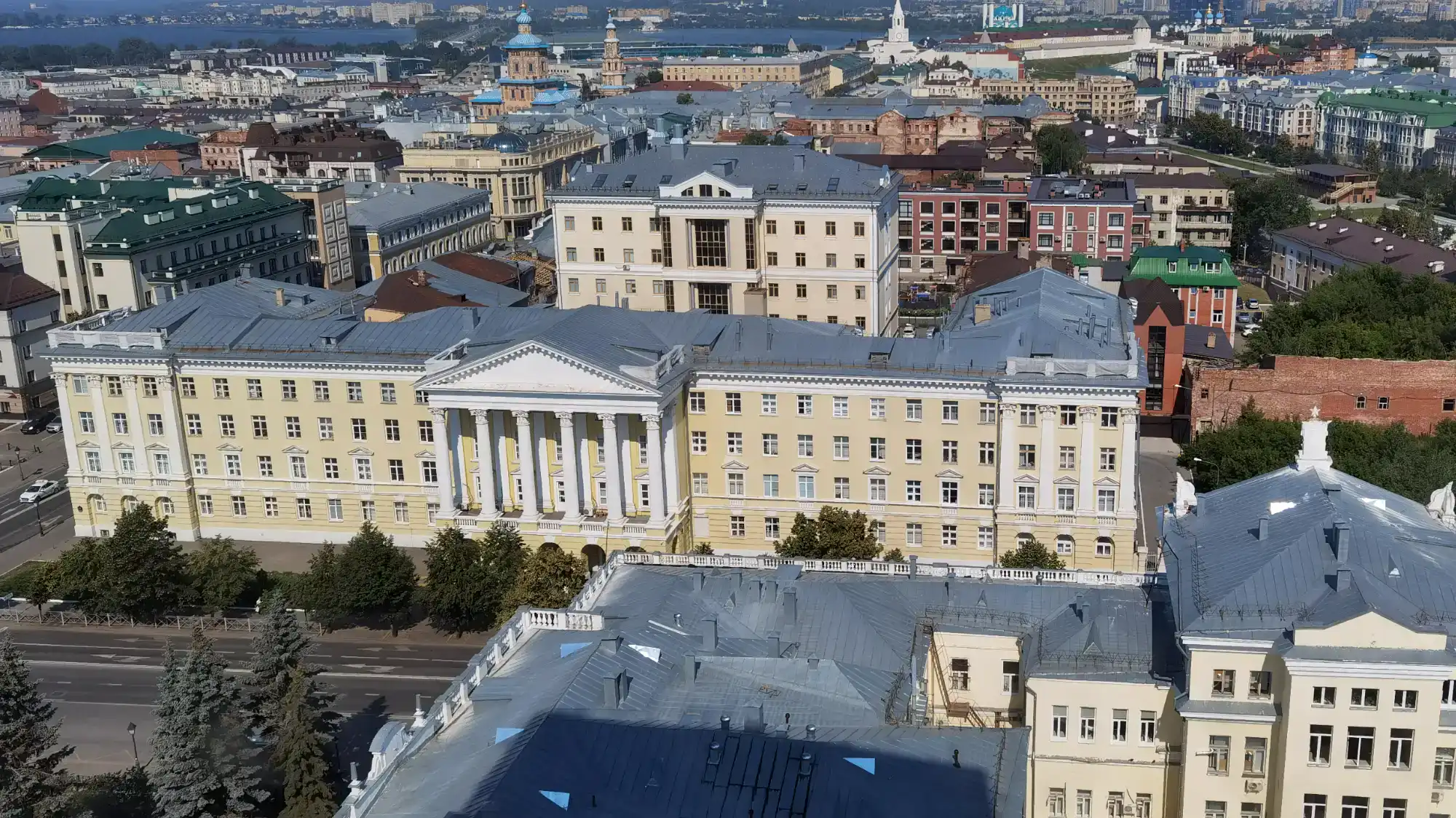 Вид сверху на исторический центр города с крупным жёлтым зданием в классическом стиле, рекой и храмами на заднем плане.