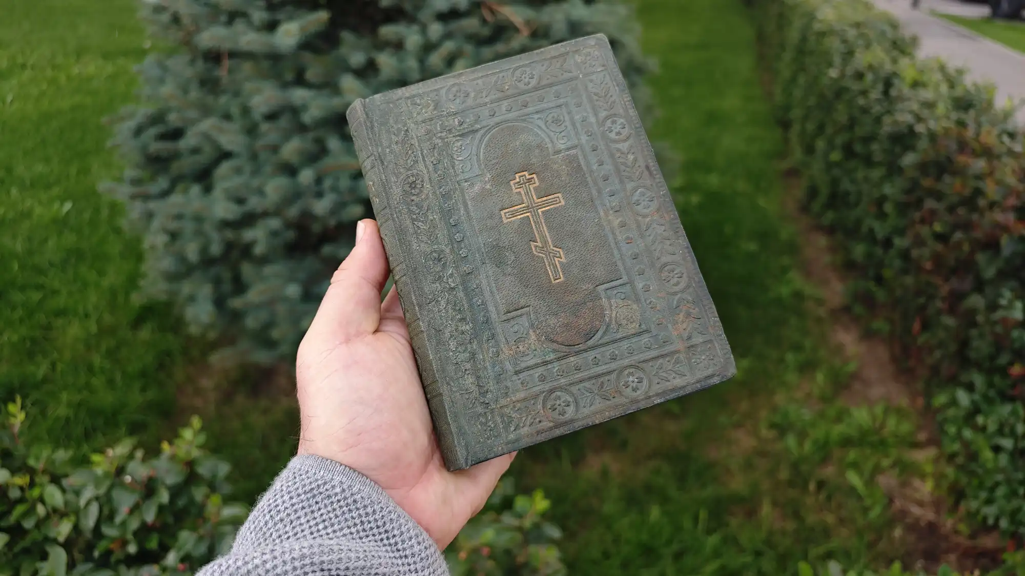 Рука в сером свитере держит старую потёртую книгу с тиснёным православным крестом на обложке, на фоне зелёной травы и кустов.