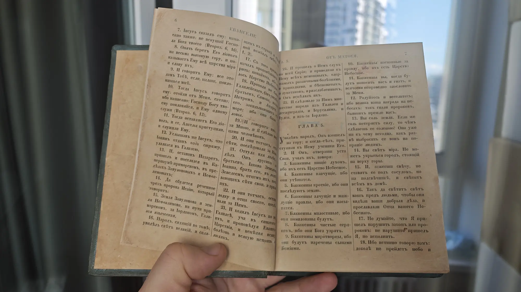 Открытая старая книга с текстом Евангелия от Матфея, глава 5, видны стихи 1–18, включая Нагорную проповедь, на фоне окна.