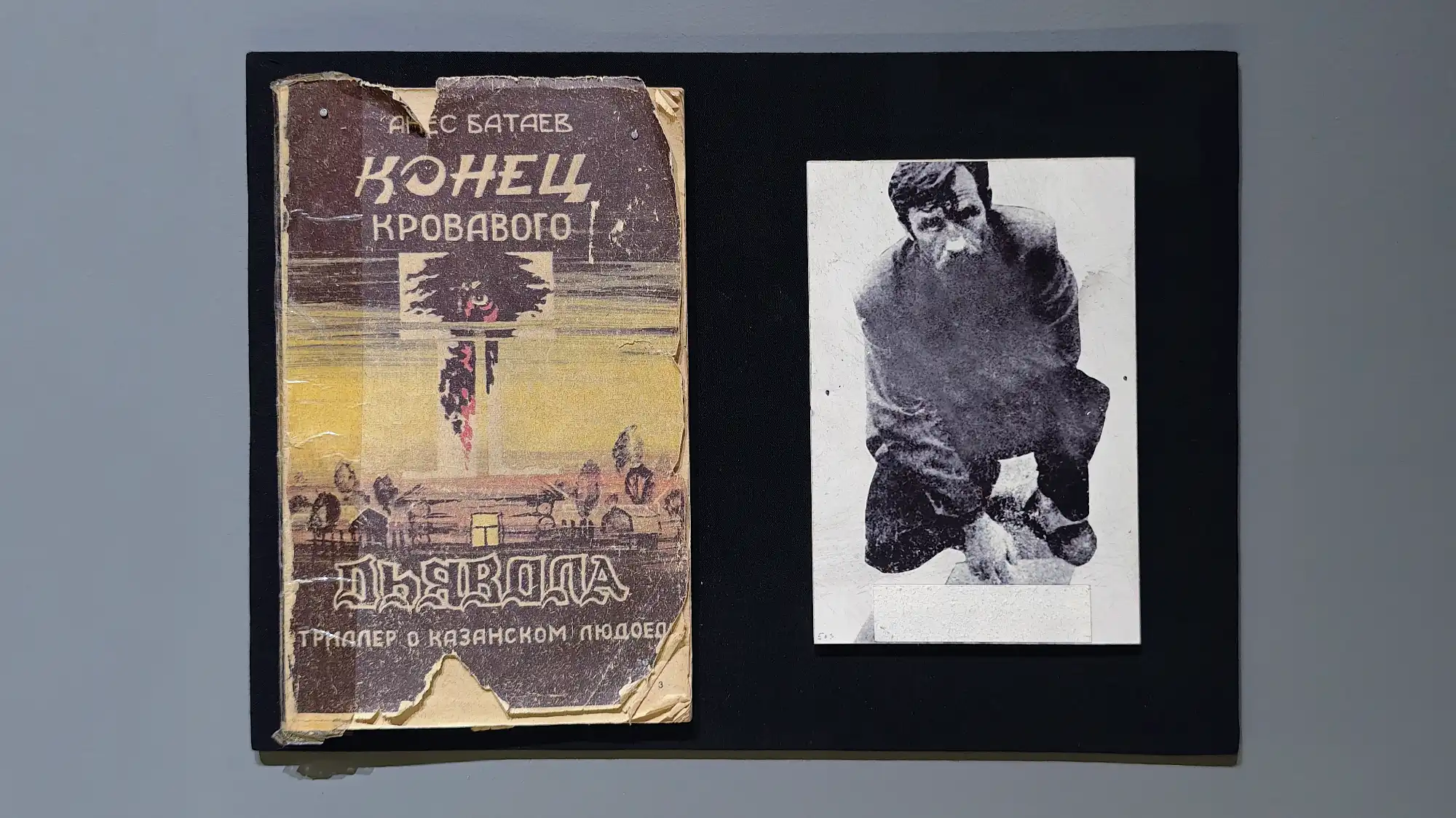 Обветшалая обложка книги Алексея Батаева «Конец кровавого дьявола. Триллер о казанском людоеде» рядом с чёрно-белой фотографией мужчины в позе на корточках.