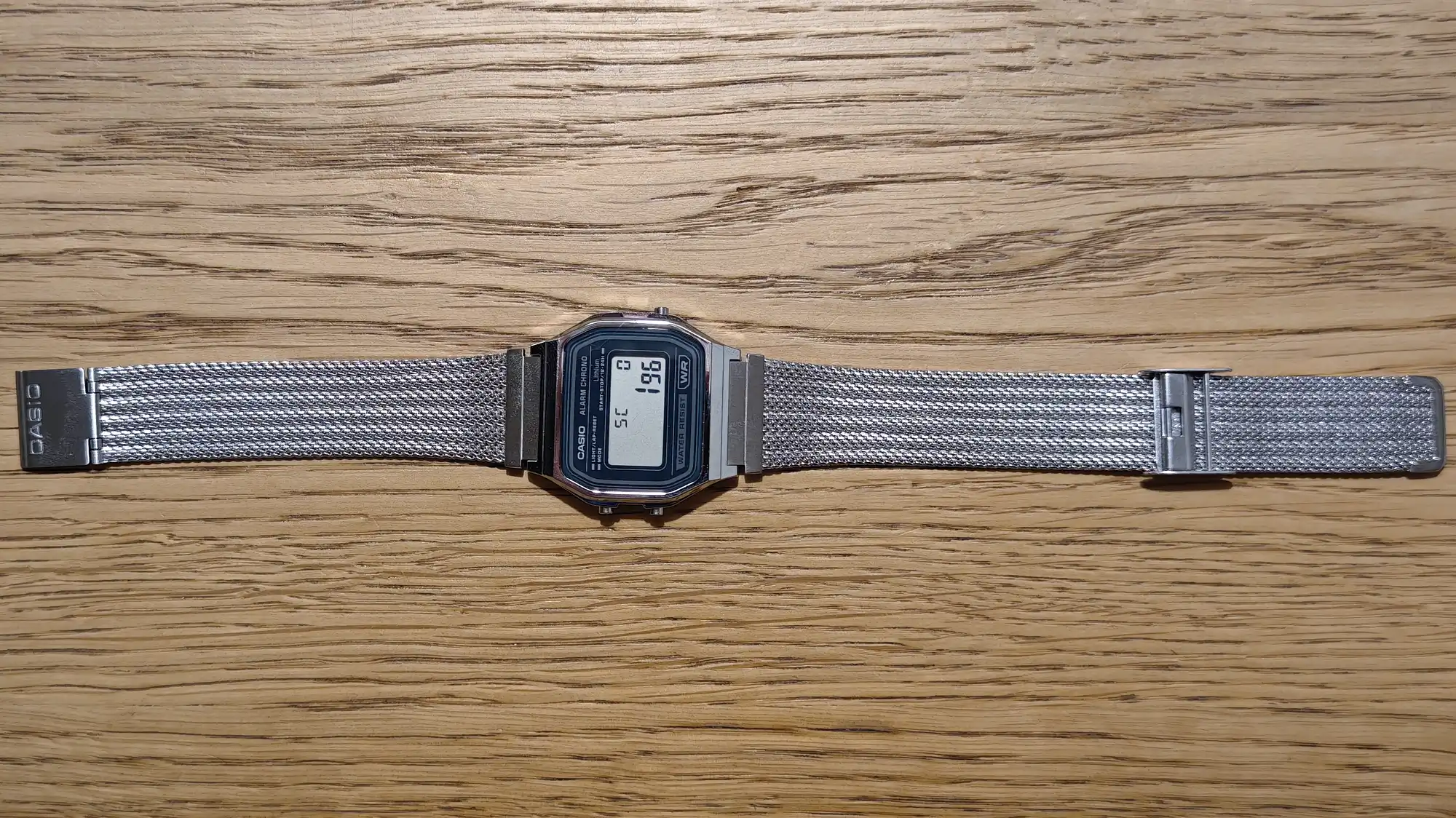 Цифровые наручные часы Casio с металлическим сетчатым ремешком на деревянном фоне; на дисплее отображается время 19:6, индикаторы SC и WR.