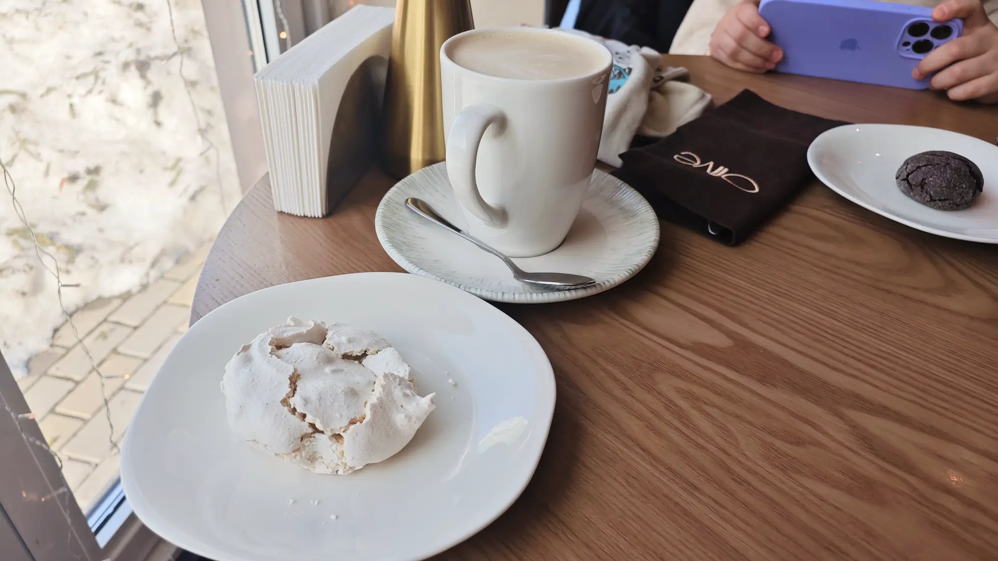 Белая чашка кофе с пенкой на блюдце, белое печенье на тарелке, шоколадное печенье на другой тарелке, руки с фиолетовым телефоном, салфетка с надписью «Olive», вид из окна на снег.