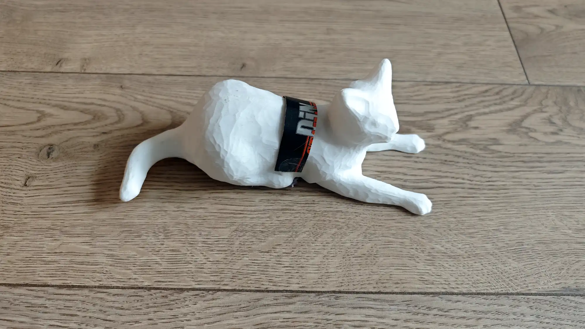 Белая 3D-модель лежащей кошки с видимыми слоями печати, на шее которой надет черный браслет с надписью «DIN» и оранжевыми полосками, на деревянном полу.