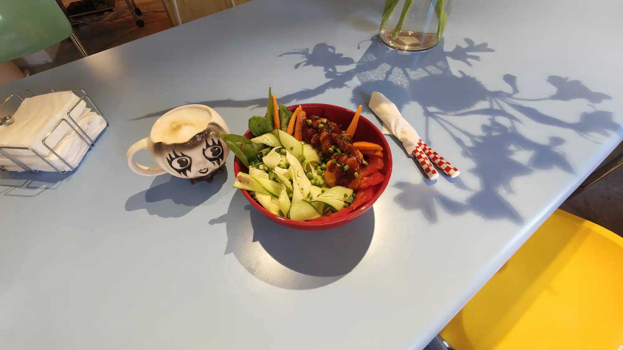 Красная миска с салатом из огурцов, моркови, горошка и мяса в соусе, рядом чашка с рисунком лица, салфетки и тень от растения на светло-голубом столе.