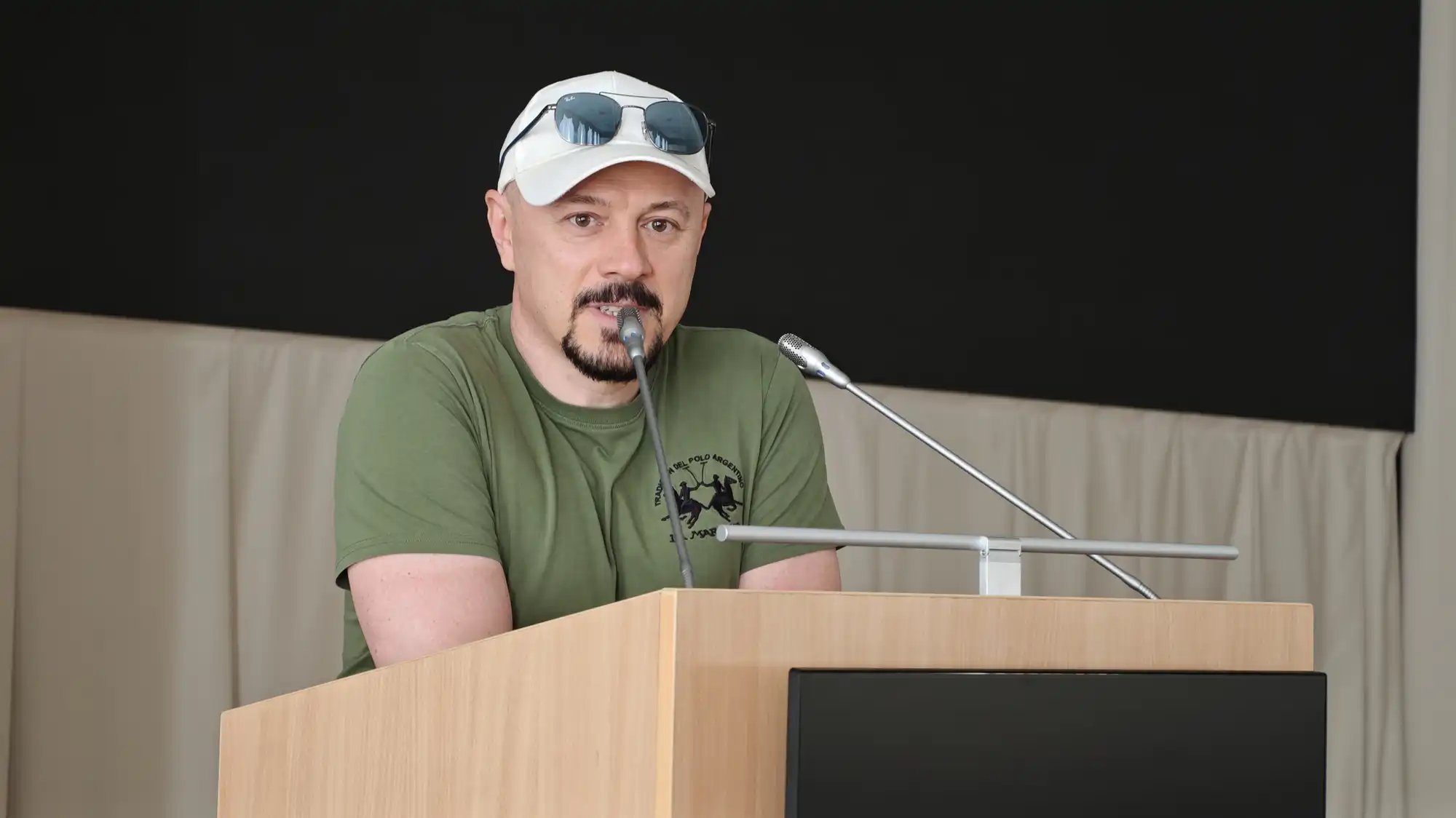 Мужчина в зелёной футболке и белой кепке с очками на козырьке стоит за деревянной трибуной и говорит в микрофон на фоне светлой шторы и чёрного экрана.