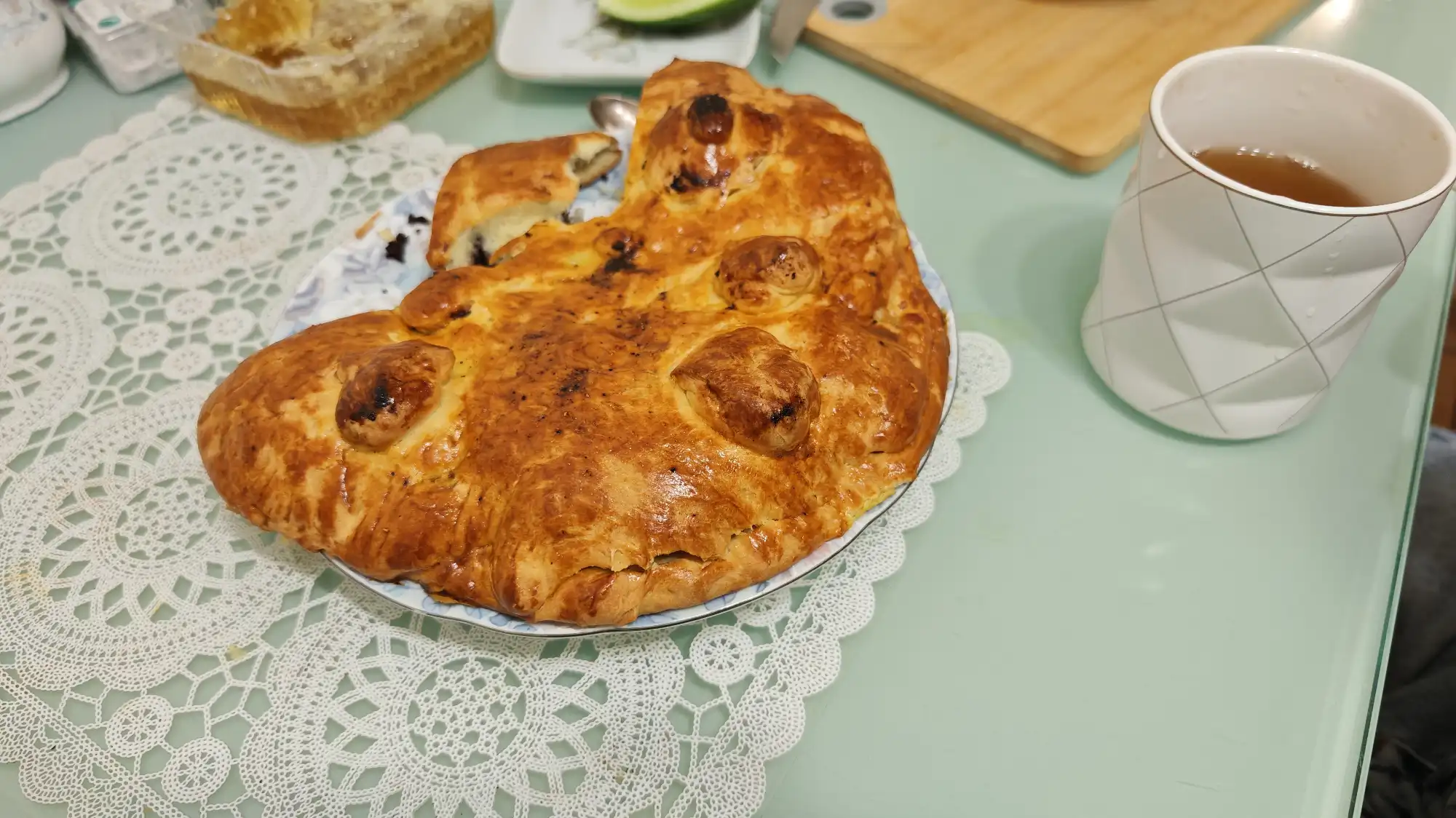 Золотистый пирог с начинкой на тарелке, рядом чашка чая и кусочек лимона на столе с кружевной салфеткой.