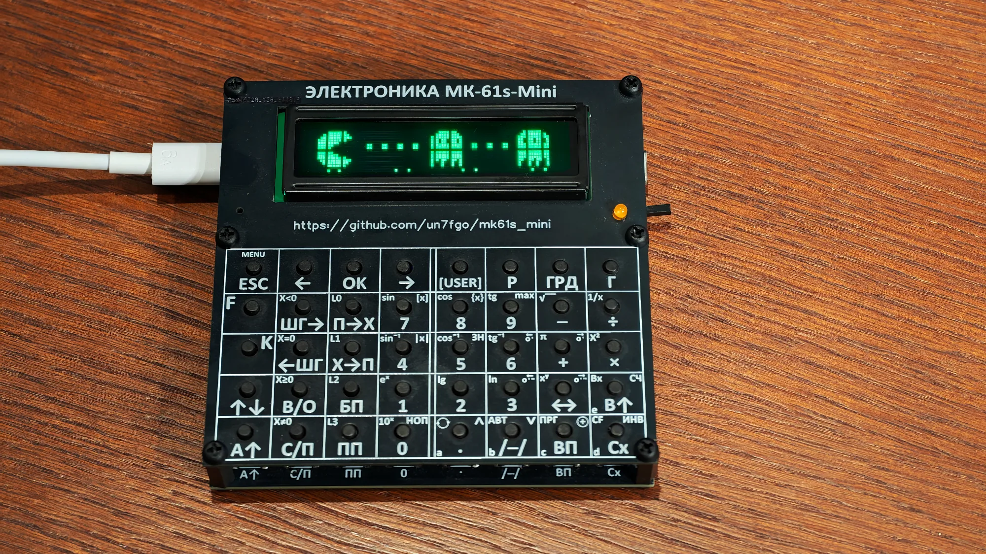 На чёрном корпусе калькулятора МК-61с Mini видны зелёные пиксельные фигурки, рядом подключён белый USB-кабель.
